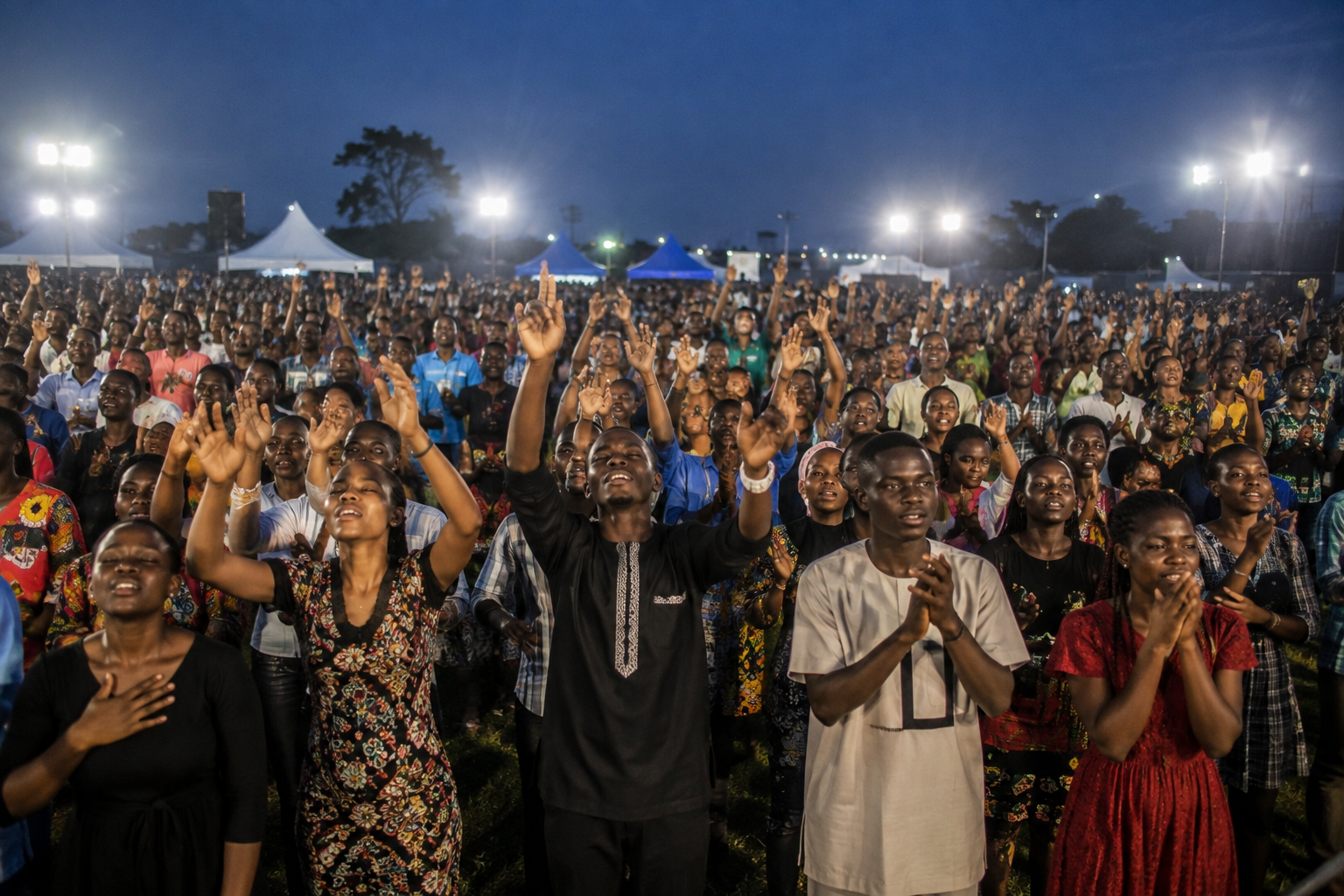 A large crowd of worshippers with raised hands at an open-air Gospel crusade at dusk